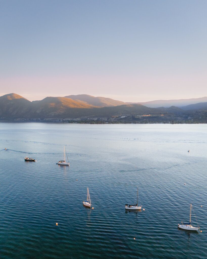 A drone image captures a summertime sunset in Naramata, British Columbia. Sailboats cover the lake as the golden sun covers the rolling hills.