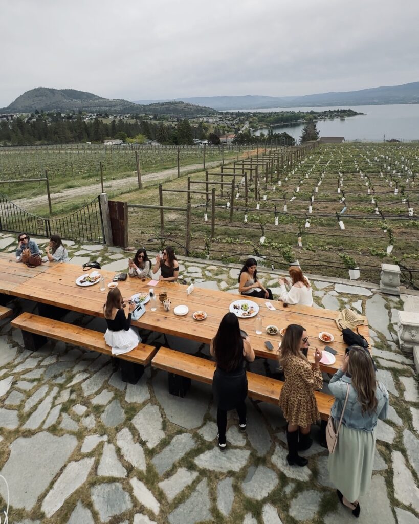 Girls sit on a long picnic bench next to an Okanagan vineyard, sipping wine and eating charcuterie in Kelowna, British Columbia