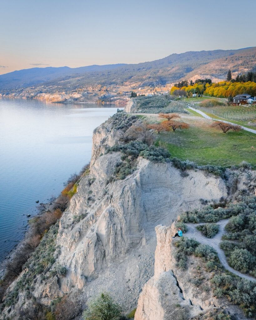 A drone image captures the towering clay cliffs of the Kettle Valley Trail near Penticton, British Columbia. A girl sits on the edge of the cliff looking out toward the valley