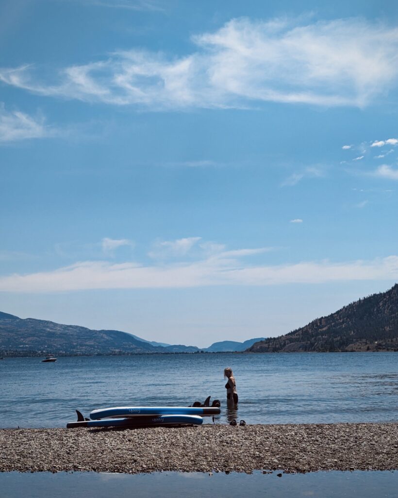 A woman stands knee deep in water next to two paddle boards on Sunoka Beach in The Okanagan Valley, British Columbia
