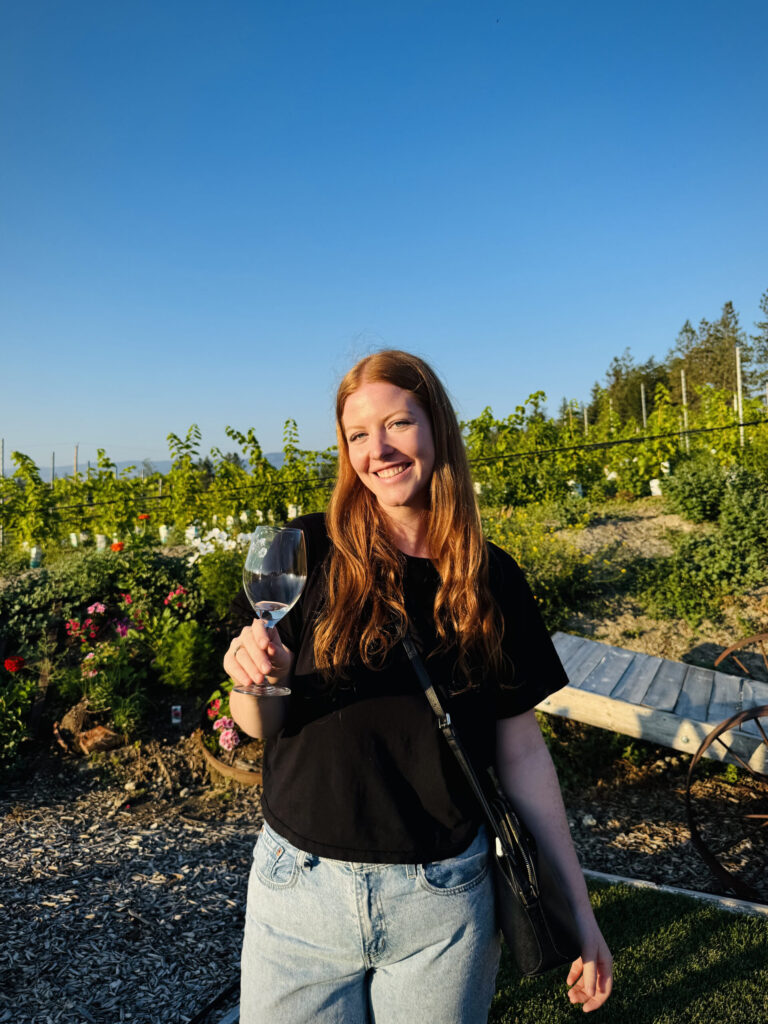 A girl stands with a wine glass in hand in front of vineyards on a sunny day in Kelowna, British Columbia